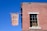 A red brick building with a rectangular window partially covered by a sign inside that reads 'DEFUND THE POLICE'. There is a brown banner hanging outside that reads 'De Stefano's Steak & Chops'. A street sign on the corner of the building displays 'CONSELYEA ST'. The sky is clear and blue.