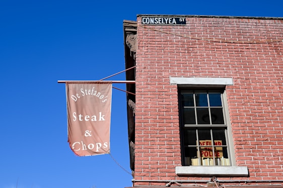 A red brick building with a rectangular window partially covered by a sign inside that reads 'DEFUND THE POLICE'. There is a brown banner hanging outside that reads 'De Stefano's Steak & Chops'. A street sign on the corner of the building displays 'CONSELYEA ST'. The sky is clear and blue.