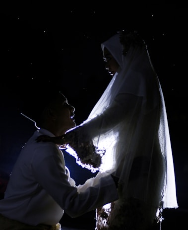 A silhouette of a couple appears against a dark background, with the woman wearing a veil and traditional attire, and the man looking up towards her. They are intimately positioned, suggesting a moment of closeness or celebration.