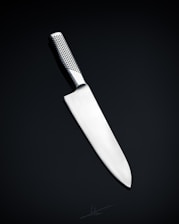A sleek, silver kitchen knife with a stainless steel blade and a textured handle is positioned against a dark, neutral background. The knife's design is modern and minimalistic, emphasizing its sharpness and craftsmanship.