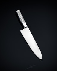 A sleek, silver kitchen knife with a stainless steel blade and a textured handle is positioned against a dark, neutral background. The knife's design is modern and minimalistic, emphasizing its sharpness and craftsmanship.