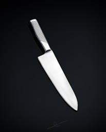 A sleek, silver kitchen knife with a stainless steel blade and a textured handle is positioned against a dark, neutral background. The knife's design is modern and minimalistic, emphasizing its sharpness and craftsmanship.