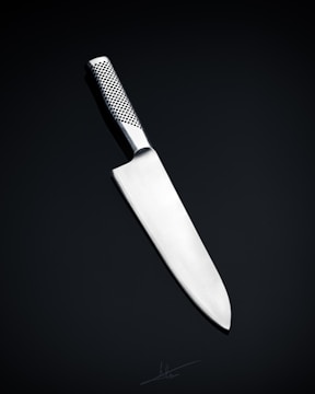 A sleek, silver kitchen knife with a stainless steel blade and a textured handle is positioned against a dark, neutral background. The knife's design is modern and minimalistic, emphasizing its sharpness and craftsmanship.