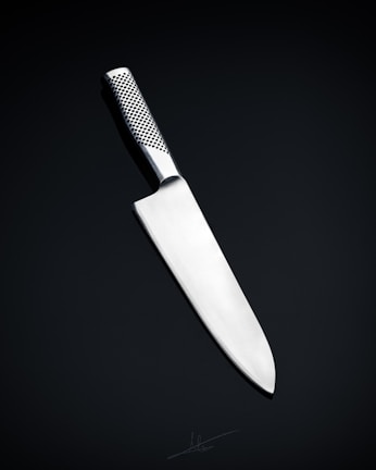 A sleek, silver kitchen knife with a stainless steel blade and a textured handle is positioned against a dark, neutral background. The knife's design is modern and minimalistic, emphasizing its sharpness and craftsmanship.