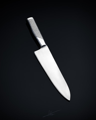 A sleek, silver kitchen knife with a stainless steel blade and a textured handle is positioned against a dark, neutral background. The knife's design is modern and minimalistic, emphasizing its sharpness and craftsmanship.