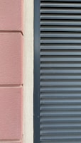 A close-up view of a building facade features a section of textured pinkish-brown plaster next to a cream-colored surface. Adjacent to these is a set of dark gray louvered shutters, creating a stark contrast between the smooth, modern surface and the textured, traditional wall.