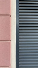 A close-up view of a building facade features a section of textured pinkish-brown plaster next to a cream-colored surface. Adjacent to these is a set of dark gray louvered shutters, creating a stark contrast between the smooth, modern surface and the textured, traditional wall.