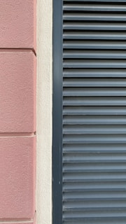 A close-up view of a building facade features a section of textured pinkish-brown plaster next to a cream-colored surface. Adjacent to these is a set of dark gray louvered shutters, creating a stark contrast between the smooth, modern surface and the textured, traditional wall.
