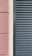A close-up view of a building facade features a section of textured pinkish-brown plaster next to a cream-colored surface. Adjacent to these is a set of dark gray louvered shutters, creating a stark contrast between the smooth, modern surface and the textured, traditional wall.