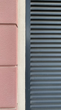 A close-up view of a building facade features a section of textured pinkish-brown plaster next to a cream-colored surface. Adjacent to these is a set of dark gray louvered shutters, creating a stark contrast between the smooth, modern surface and the textured, traditional wall.