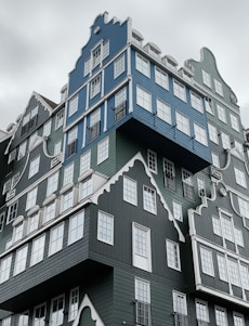 A modern, uniquely designed building composed of stacked houses with gabled roofs painted in shades of blue and green. The structure features numerous white-framed windows, creating a whimsical and intricate appearance.