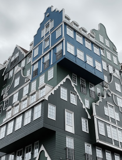 A modern, uniquely designed building composed of stacked houses with gabled roofs painted in shades of blue and green. The structure features numerous white-framed windows, creating a whimsical and intricate appearance.