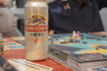 A can of Kirin Ichiban beer is placed prominently next to a board game setup on a table. The can displays a vibrant design featuring a mythical creature. In the background, board game pieces and stacks of cards are spread across a colorful game board. A person sits nearby, partially visible.