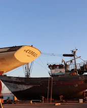 Several boats are under maintenance in a shipyard with clear blue skies overhead. The prominent boat in the foreground is labeled 'CYPR.AE.' and resting on supports, with visible ladders and equipment nearby. Other boats are visible in the background, all positioned on a concrete surface near the water.