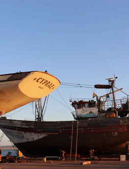 Several boats are under maintenance in a shipyard with clear blue skies overhead. The prominent boat in the foreground is labeled 'CYPR.AE.' and resting on supports, with visible ladders and equipment nearby. Other boats are visible in the background, all positioned on a concrete surface near the water.