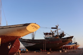 Several boats are under maintenance in a shipyard with clear blue skies overhead. The prominent boat in the foreground is labeled 'CYPR.AE.' and resting on supports, with visible ladders and equipment nearby. Other boats are visible in the background, all positioned on a concrete surface near the water.