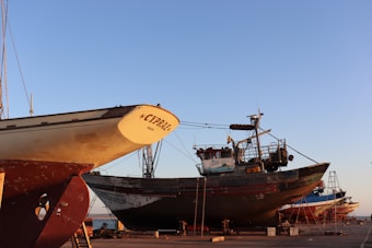 Several boats are under maintenance in a shipyard with clear blue skies overhead. The prominent boat in the foreground is labeled 'CYPR.AE.' and resting on supports, with visible ladders and equipment nearby. Other boats are visible in the background, all positioned on a concrete surface near the water.