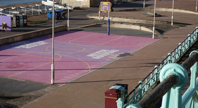 A vibrant outdoor basketball court with a pink surface, yellow lines, and a purple basketball hoop. The court is situated along a promenade with railings, bins, and a beach area nearby. The setting appears to be quiet with very few people in the vicinity.