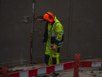 A person, dressed in high-visibility work attire with a bright orange hood, is standing near a red and white barrier in a construction zone. The individual is wearing ear protection and appears to be working in a wet or muddy environment, with some dirt visible on their clothing.