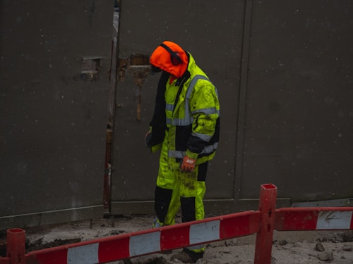 A person, dressed in high-visibility work attire with a bright orange hood, is standing near a red and white barrier in a construction zone. The individual is wearing ear protection and appears to be working in a wet or muddy environment, with some dirt visible on their clothing.
