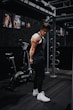 A fit individual in a black tank top and pants is standing in a gym beside a fitness bike. The person is positioned near gym equipment, including metal bars and exercise machines, and appears to be in a pensive or introspective pose. The gym walls are adorned with posters of muscular figures.