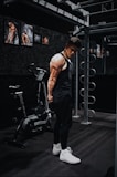 A fit individual in a black tank top and pants is standing in a gym beside a fitness bike. The person is positioned near gym equipment, including metal bars and exercise machines, and appears to be in a pensive or introspective pose. The gym walls are adorned with posters of muscular figures.