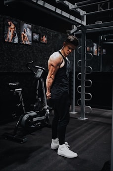 A fit individual in a black tank top and pants is standing in a gym beside a fitness bike. The person is positioned near gym equipment, including metal bars and exercise machines, and appears to be in a pensive or introspective pose. The gym walls are adorned with posters of muscular figures.