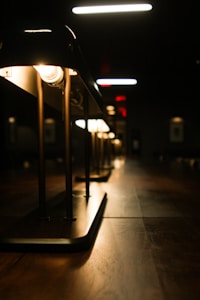 A row of elegant lamps is illuminated against a dark background, casting a warm glow on a wooden surface. The perspective of the shot gives depth, highlighting the sleek design and symmetry of the lamp fixtures.
