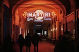 A group of people walk under an archway with warm lighting. A brightly lit Martini sign with a yellow circle and neon orange elements highlights the entrance of a cafe named 'Caffè Ristorante Torino'. The setting has an inviting, cozy atmosphere.