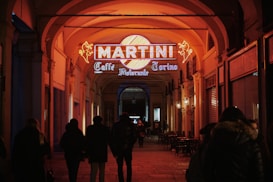 A group of people walk under an archway with warm lighting. A brightly lit Martini sign with a yellow circle and neon orange elements highlights the entrance of a cafe named 'Caffè Ristorante Torino'. The setting has an inviting, cozy atmosphere.