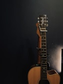 An acoustic guitar is propped against a dark wall, partially illuminated by soft lighting from the right side. The guitar has a natural wood finish, and its tuning pegs are visible. The overall environment appears calm and focused on the instrument.
