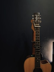An acoustic guitar is propped against a dark wall, partially illuminated by soft lighting from the right side. The guitar has a natural wood finish, and its tuning pegs are visible. The overall environment appears calm and focused on the instrument.