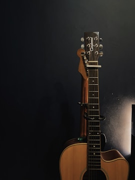 An acoustic guitar is propped against a dark wall, partially illuminated by soft lighting from the right side. The guitar has a natural wood finish, and its tuning pegs are visible. The overall environment appears calm and focused on the instrument.