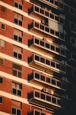 A multi-story apartment building facade with a series of balconies aligned vertically. Red brick and white paneling dominate the exterior, with each balcony equipped with glass railings. Some balconies feature air conditioning units and awnings, while sunlight casts shadows against the building, creating a pattern of light and shadow.