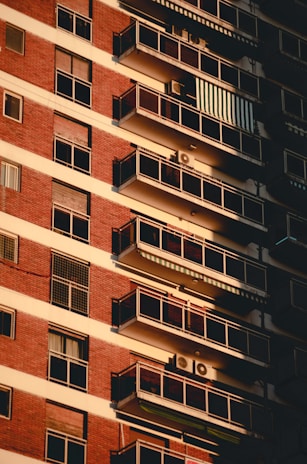 A multi-story apartment building facade with a series of balconies aligned vertically. Red brick and white paneling dominate the exterior, with each balcony equipped with glass railings. Some balconies feature air conditioning units and awnings, while sunlight casts shadows against the building, creating a pattern of light and shadow.