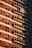 A multi-story apartment building facade with a series of balconies aligned vertically. Red brick and white paneling dominate the exterior, with each balcony equipped with glass railings. Some balconies feature air conditioning units and awnings, while sunlight casts shadows against the building, creating a pattern of light and shadow.