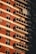 A multi-story apartment building facade with a series of balconies aligned vertically. Red brick and white paneling dominate the exterior, with each balcony equipped with glass railings. Some balconies feature air conditioning units and awnings, while sunlight casts shadows against the building, creating a pattern of light and shadow.