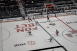 A hockey game in progress on an ice rink, with players wearing red and white jerseys. A referee is present, and there are advertisements surrounding the rink. The stands partially filled with spectators suggest a lively yet contained atmosphere.
