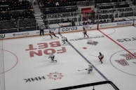 A hockey game in progress on an ice rink, with players wearing red and white jerseys. A referee is present, and there are advertisements surrounding the rink. The stands partially filled with spectators suggest a lively yet contained atmosphere.