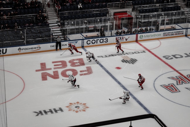A hockey game in progress on an ice rink, with players wearing red and white jerseys. A referee is present, and there are advertisements surrounding the rink. The stands partially filled with spectators suggest a lively yet contained atmosphere.