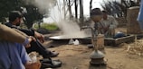 A rural outdoor setting with a traditional hookah prominently placed in the foreground. Two men are sitting near a cooking area where steam rises, suggesting active cooking or brewing. The background shows trees and an open field under a partly cloudy sky.