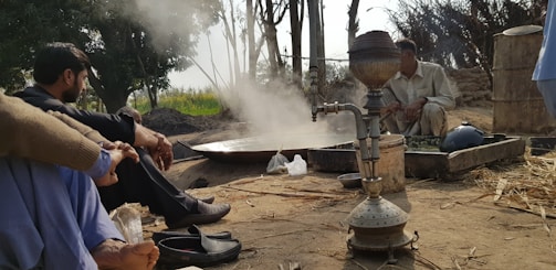A rural outdoor setting with a traditional hookah prominently placed in the foreground. Two men are sitting near a cooking area where steam rises, suggesting active cooking or brewing. The background shows trees and an open field under a partly cloudy sky.