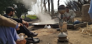 A rural outdoor setting with a traditional hookah prominently placed in the foreground. Two men are sitting near a cooking area where steam rises, suggesting active cooking or brewing. The background shows trees and an open field under a partly cloudy sky.