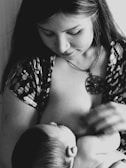 A woman is breastfeeding a baby, cradling the child close. The image is in black and white, highlighting the peaceful and intimate moment. She has long hair and is wearing a patterned top with a pendant necklace.