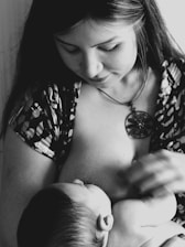A woman is breastfeeding a baby, cradling the child close. The image is in black and white, highlighting the peaceful and intimate moment. She has long hair and is wearing a patterned top with a pendant necklace.