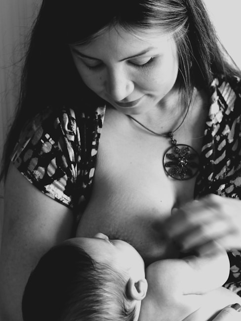 A woman is breastfeeding a baby, cradling the child close. The image is in black and white, highlighting the peaceful and intimate moment. She has long hair and is wearing a patterned top with a pendant necklace.
