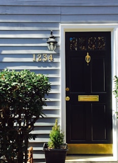 A black door with a golden mail slot and door knocker is set against a light blue wooden paneled wall. The house number, 1234, is displayed prominently in gold. A classic outdoor lantern is mounted on the wall next to the door. Green shrubs and a small potted plant decorate the entrance.