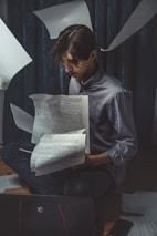 A person sits on the floor surrounded by floating papers, appearing deep in concentration as they look at documents in their hands. The setting is dimly lit, conveying a focused and studious atmosphere. A laptop is visible in the foreground, and the curtains in the background add to the moody setting.