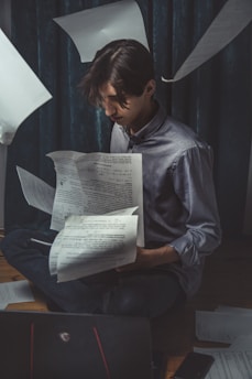 A person sits on the floor surrounded by floating papers, appearing deep in concentration as they look at documents in their hands. The setting is dimly lit, conveying a focused and studious atmosphere. A laptop is visible in the foreground, and the curtains in the background add to the moody setting.