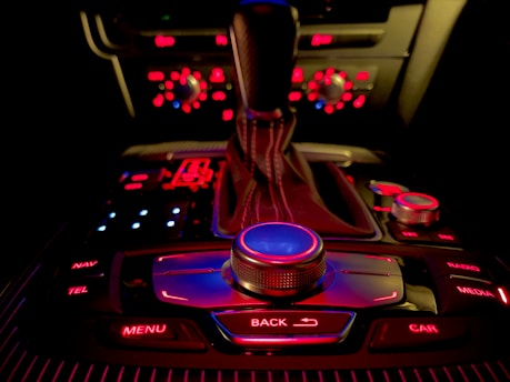 A car's central console with a gear shift lever and various buttons and knobs, all illuminated with red and blue backlighting. The buttons include options for navigation, media, telephone, menu, car, and more.